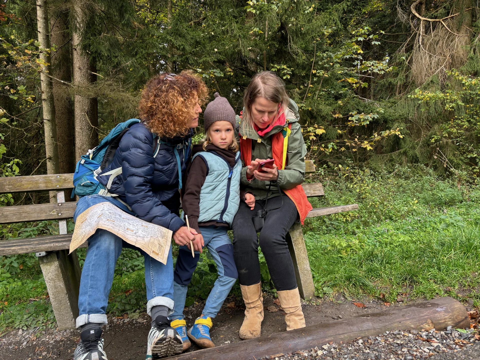 2 Frauen und 1 Kind sitzen auf einer Bank und lesen die Schatzkarte und nutzen das Smartphone